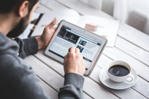 Alcance real: como transformar um simples post em matéria de portal A man uses a tablet to browse news while enjoying a cup of coffee, stylus in hand.